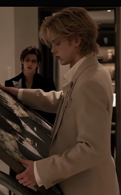The hung picture — a light-blond man in a pale-apricot suit steadying a dark floral painting against the wall, a dark-haired man in a black suit with a grey-trimmed collar watching him close from behind (ep2; atmosphere beat — watched-while-working frame)