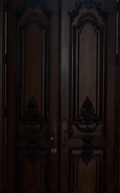 The doors - tall carved double doors of dark walnut with aged brass levers and ornate plates in a dim warm hall (ep6; scene beat - the estate interiors, no figures)