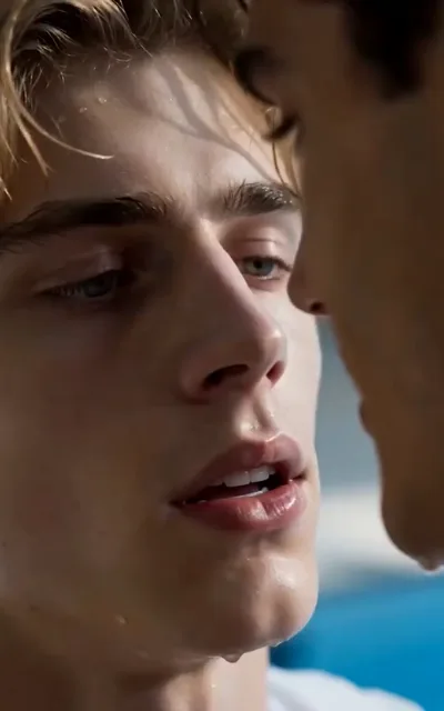 The water-lit gaze — close-up of a light golden-brown-haired young man soaked from the pool, fine droplets across his brow and jaw, a hanging drop at his chin, half-lidded eyes and parted lips under striped sunlight, another man's darker water-beaded profile a breath away at frame right (ep1; intimacy-adjacent frame as aired, not amplified)