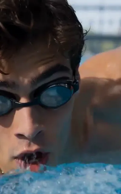 The breath — a dark-curled swimmer in black-framed race goggles half-submerged at the water line, mouth open exhaling a fine spray of bubbles and droplets, bright shallow pool water below with sunlit ripples beyond (ep1; atmosphere beat — training-breath frame)