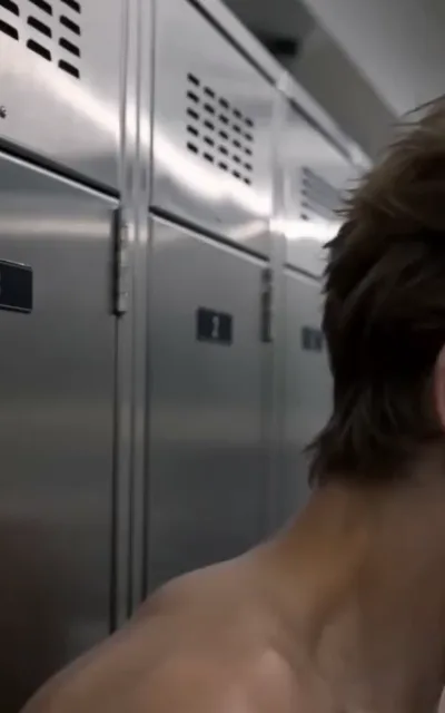 The locker row — the back of a dark-curled head and bare shoulders at frame right facing a long row of steel lockers with vent slits and numbered black plates, a clear number 2 on the middle door, the corridor receding in soft focus (ep3; atmosphere beat — changing-room frame)