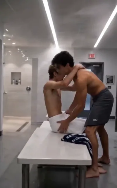 The shower-room bench — in a bright tiled shower room with LED light strips, a slim light golden-brown-haired man in a white bath towel seated on the bench, arms raised around the neck of a taller dark-curled man in grey shorts leaning in close over him, both bare-chested (ep4; intimacy-adjacent frame as aired, not amplified)