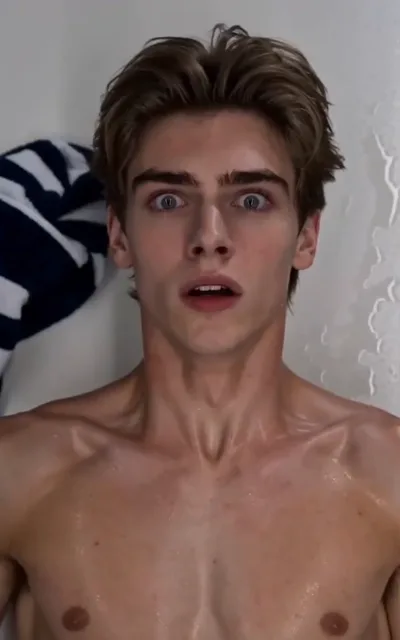 The shower shock — a light golden-brown-haired young man under the spray, skin beaded with water, blue-grey eyes wide and mouth open in plain astonishment, a navy-and-white striped towel at frame corner, runnels on the white tile behind (ep4; atmosphere beat — caught-off-guard frame)