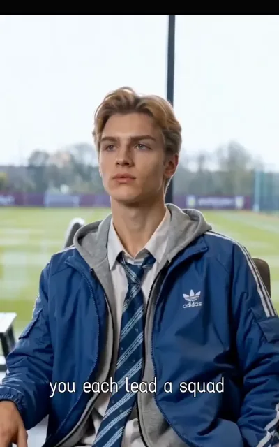 The locker-room chalk talk — the coach's board and the squad leaning in, one player in a royal-blue kit with a clover-shaped crest on the chest pointing at the pitch map, the caption “you each lead a squad” beneath (ep1; subtitles as aired; brand-pattern kept as aired — clover-shaped crest as broadcast, described generically, no brand name in this copy)