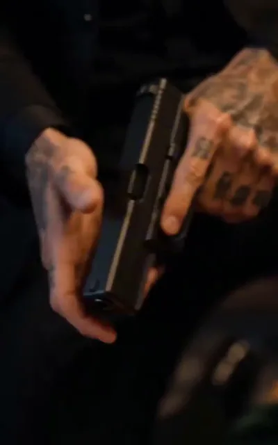 Frame ep05-f01 from episode 5 — A loaded gun in tattooed hands (ep5; subtitles as aired — The hands holding the gun are covered in ink.)