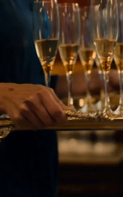 Frame ep01-f00 from episode 1 — Lucia carries a tray of champagne flutes through the dark hall (ep1; subtitles as aired — Dante Falcon is here for me tonight.)