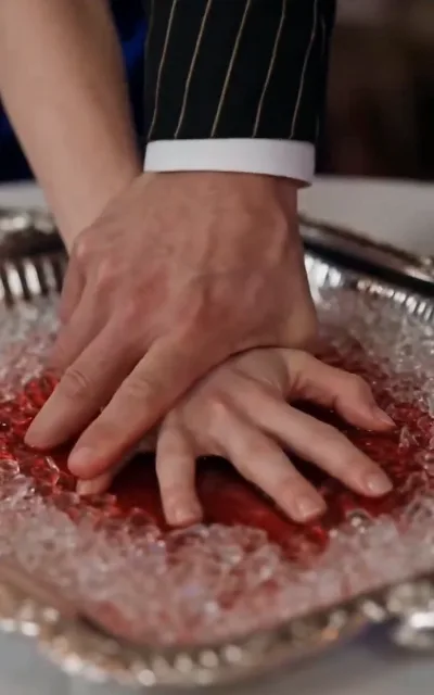 An adult hand pressing a child's hand onto a tray of ice and red liquid — the organ-trade scheme in one image (ep6)
