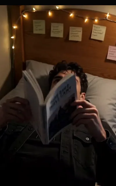 Liam half-propped on his pillow reading, the book hiding most of his face — string lights and handwritten notes on the headboard behind him, one reading "That must hurt" (ep5)