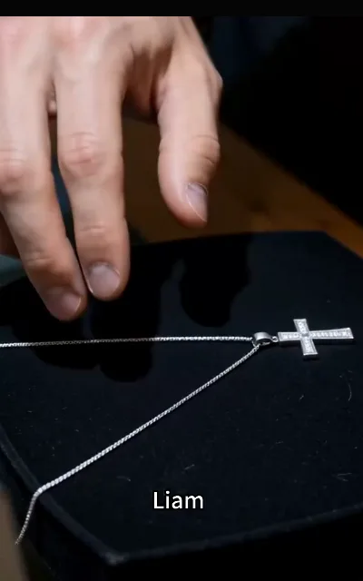 A silver chain with a stone-set cross pendant laid on black velvet, a hand reaching toward it — the station’s own caption at the bottom of the frame reads "Liam", a design element presented as shown and not further anchored (ep6, platform annotation as aired)