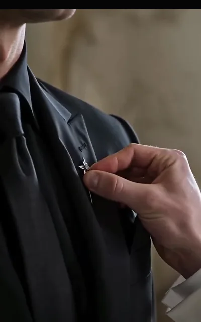 Eli fastening a star-shaped lapel pin onto Kalen’s black formal wear, hands close and careful — the last quiet intimacy before the vows (ep1)