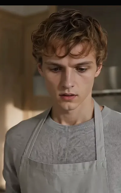 Eli in knit and apron, head lowered mid-task in the kitchen — the giving years in one domestic frame (ep2)