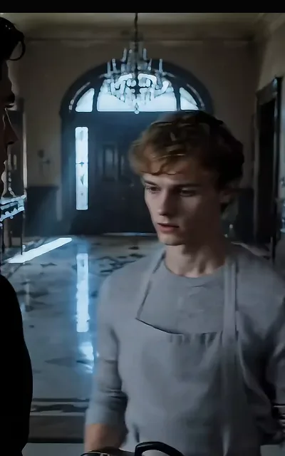 The mansion’s long corridor: Eli carrying his day’s work past a dark-haired figure in profile — the household axis between the two of them, staged exactly as shot (ep2)