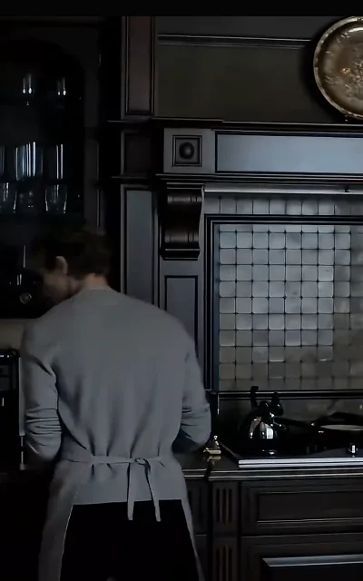 Eli from behind at the kitchen work — cardigan, kettle, silverware — a marriage kept running by one pair of hands (ep2)