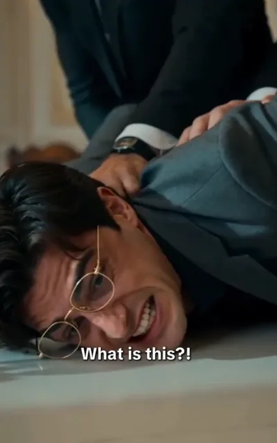 The challenge - a man with dark hair and fine gold round frames in a grey suit seized and held back mid-outburst in a bright formal room, the caption What is this?! beneath (ep1; subtitles as aired - the suppressed-man conflict beat; styling-collision figure sharing the male lead's round-frame look, identity not anchored)