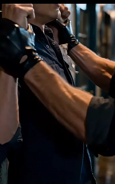 Fingerless leather gloves seizing the collar of a navy work shirt, close up — the rodeo world's casual authority over its stable hands, delivered in a single grip (ep1)