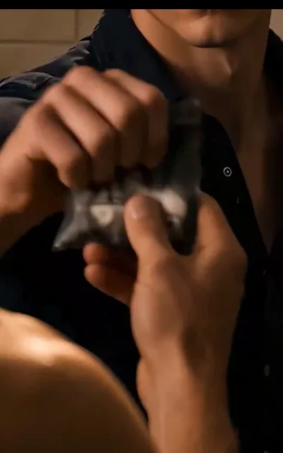 The sealed packet torn open, white blocks inside, hands and shoulders tangled at the frame's edge — the suppressant ritual and what it costs, staged as aired (ep6; tension frame as aired)
