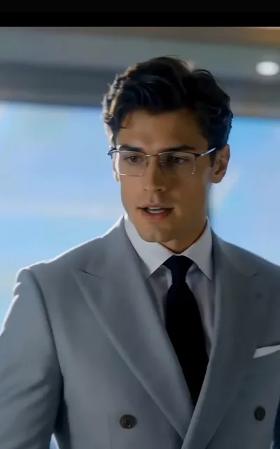 The formal portrait — a man with metal-rimmed glasses and a deep-brown undercut in a light-grey double-breasted suit, city glass behind (ep1; atmosphere beat — avatar source frame, avatar_oliver crop (0,40,300,640))