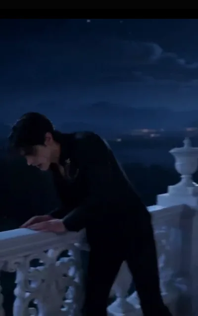 The balustrade at night — a lone figure in slim black leaning over the carved white stone rail, head bowed toward the town lights below, stars faint in the deep-blue sky (ep3; atmosphere beat)