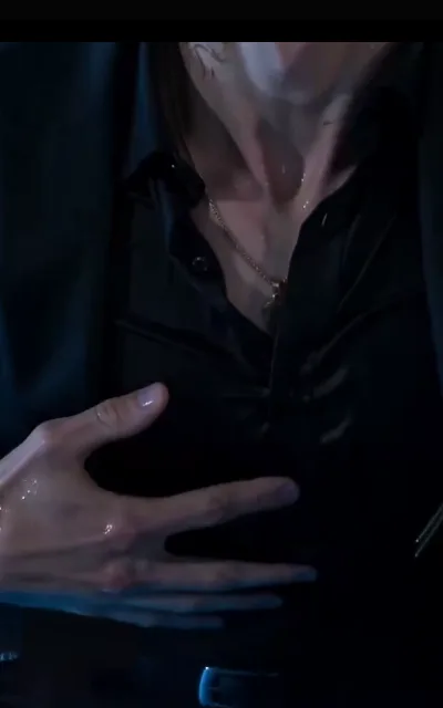 The composure study — a black open collar beaded with water, a fine gold chain, and a long-fingered hand pressed flat against the dark shirt front, silver buckle just visible at the frame edge (ep4; intimacy-adjacent frame as aired, not amplified — torso study, no second person; REPLACEMENT slot for the dropped borrowed-image ep4 batch)