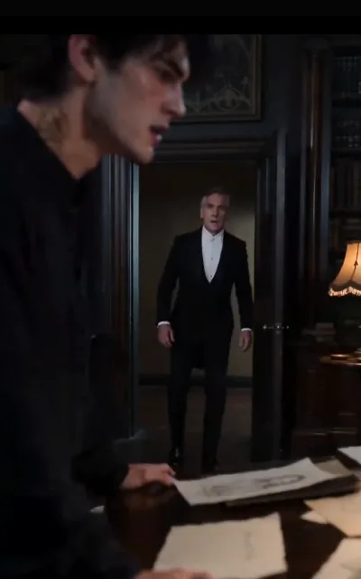 The interrogation — a young man in black at the dark wood table, yellowed papers with drawn marks spread before him, a pale gold mark behind his ear; in the doorway a white-haired older man in formal black, just arrived, demanding answers (ep8; atmosphere beat; unnamed elder, no avatar)