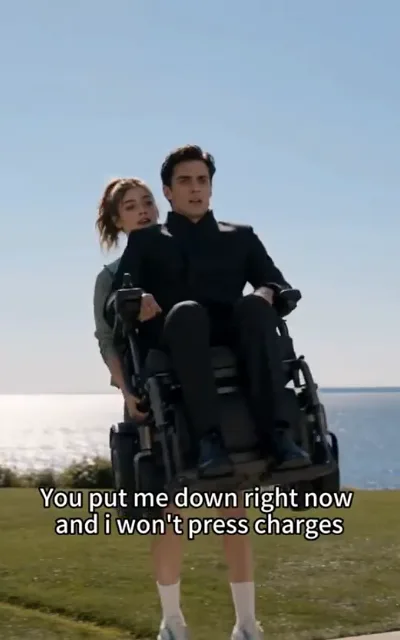 The lift — on a seafront lawn, a young woman in sporty casual wear hoisting a powered wheelchair one-handed, the man in the black stand-collar suit seated in it calling out in indignation, wide lawn and sea haze behind, the caption You put me down right now and i won't press charges beneath (ep1; subtitles as aired — TOP quote, lowercase i as-aired; HERO frame; wheelchair-context frame as aired, not amplified)