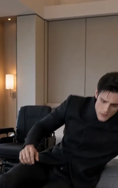 The man at ease — the male lead in a black stand-collar suit leaning back on the edge of a bed, the powered wheelchair resting in the background of the room (ep4; atmosphere beat — wheelchair-context frame as aired, not amplified)