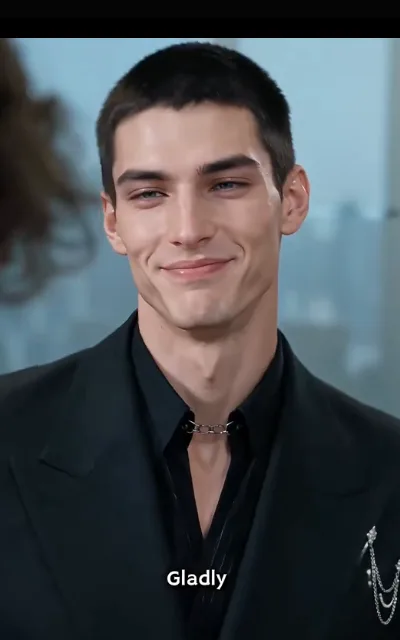 Dante in full black — suit and open collar joined by a silver chain, chained lapel brooch, a mild smirk straight at the lens — the station caption "Gladly" kept as aired (ep3; source of avatar_dante, single-base anchor; station caption as aired in frame, NOT a quote card)