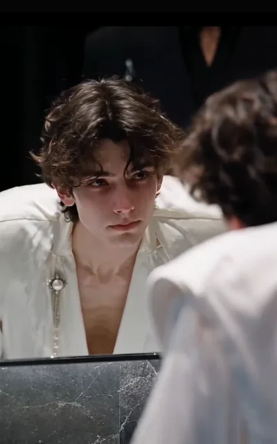 The mirror take — Elias reflected clear while his own shoulder blurs in the foreground, pearl pin at the chest, marble counter below, a dark figure lost in the background (ep5; REPLACEMENT frame for the dropped borrowed-image ep5-f15, mirror-reflection form)