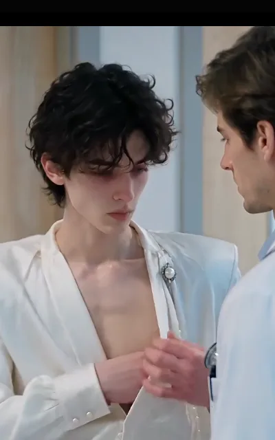 The doctor’s hands at his collar — stethoscope at the sleeve, straightening the ivory jacket for him, Elias looking down at nothing (ep6; atmosphere beat; indirect presentation, clinic consult)