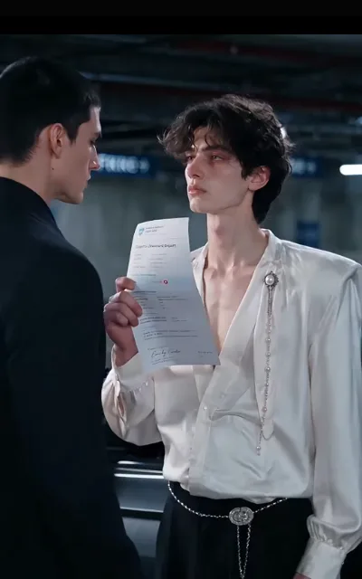 The garage standoff — Dante in black at one side, jaw set, a small mole under the ear; Elias in ivory with the printed report held up between them, chin high (ep6; HERO SOURCE)
