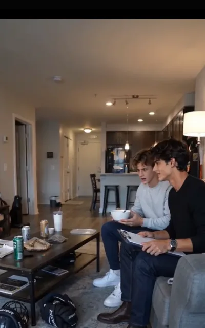 The couch stretch — her brother with a bowl and Grant with a writing board, both looking toward the camera while gear waits on the rug (ep5)