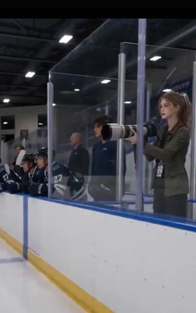 The bench view — a helmeted player in navy at the rink boards while Piper raises her long lens from the other side of the glass (ep6)