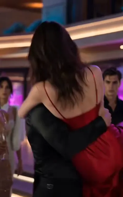 The masked embrace — in a warm-lit night lounge with pink-violet ambient glow, a man in black wrapping his arm around the waist of a woman in a red backless slip dress, his face hidden behind her, a woman in white and a man in dark wear looking on stunned (ep1; masked-mogul line beat — the passionate-night setup, identities unnamed)