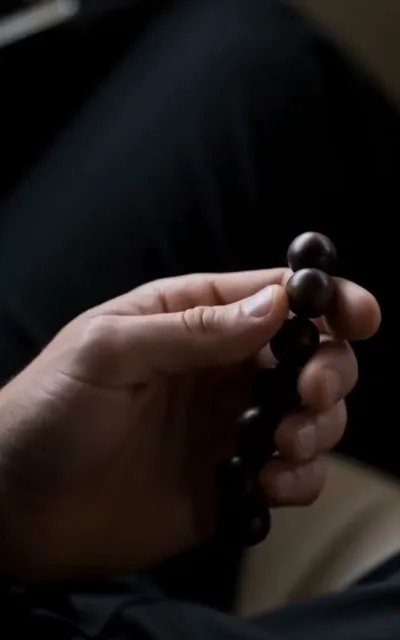 The token — in dim close focus, a man's fingers rolling a strand of dark wooden beads, two polished beads sharp against the blurred dark fabric and a pale seat edge behind (ep6; identity-token beat — the wooden bead close-up, the masked man's keepsake)