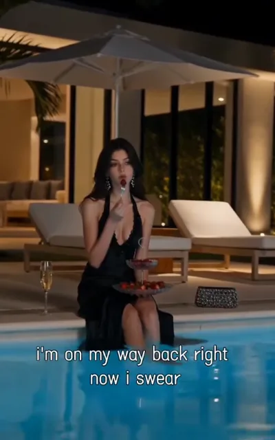 The lounge — at a night pool terrace of a warm-lit villa, a woman with dark long waves, drop diamond earrings and a black deep-V halter gown with a slit, legs dipped in the glowing water, a strawberry on a fork at her lips, a two-tier strawberry stand, a flute of sparkling wine and a crystal clutch beside, the captioned line i'm on my way back right now i swear beneath (ep8; intimacy-adjacent frame as aired, not amplified — the pool-party beat)