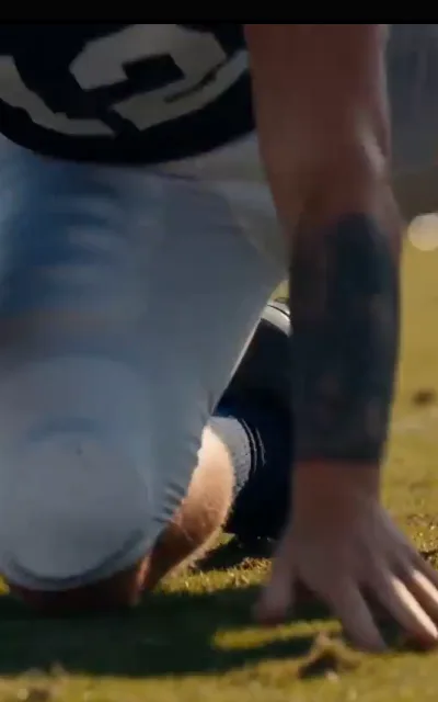 Down in the turf — a tattooed forearm planted on the grass, knee down, black jersey hem above, the set position of a play about to start (ep1; atmosphere beat)