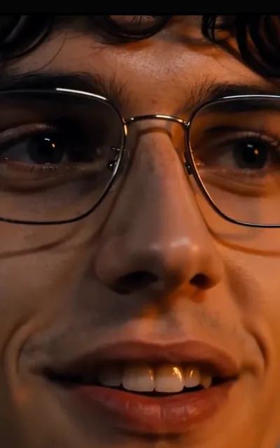 The stiff smile — glasses catching warm light, eyes wide, the corner of the mouth pulled into something between a grin and a flinch (ep2; atmosphere beat)