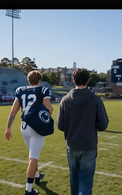 Side by side off the field — the quarterback in stripes and the grey-hoodied roommate walking the marked grass together, stands and campus tower behind (ep6; atmosphere beat)