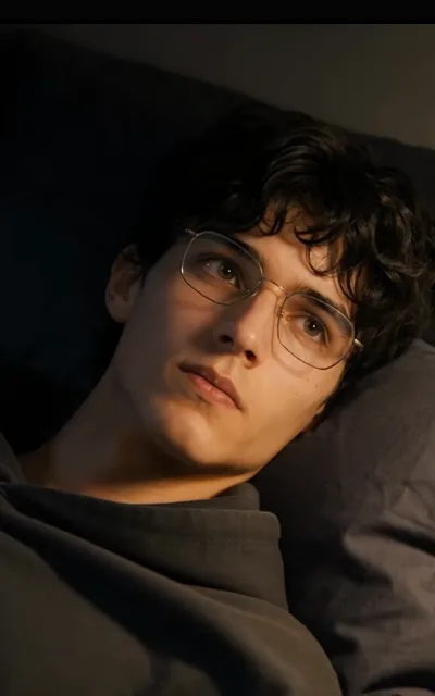 Eli at rest — glasses on, curls against the pillow, eyes drifting up and away, warm side-light (ep8; source of avatar_eli, single-base anchor)