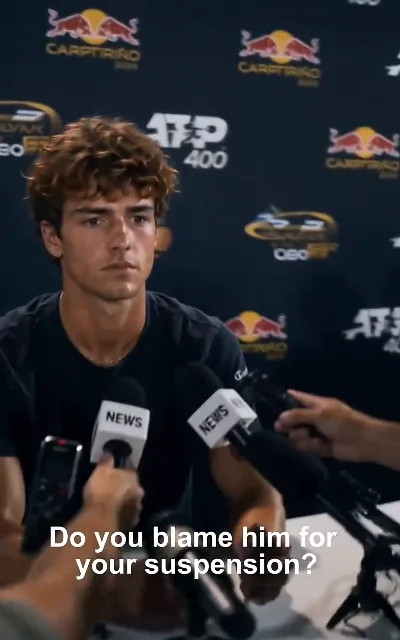 "Do you blame him for your suspension?" — Jayce at the press table in black, a tour event backdrop behind him: the question he cannot answer cleanly (ep2, subtitles as aired; backdrop boards as aired)
