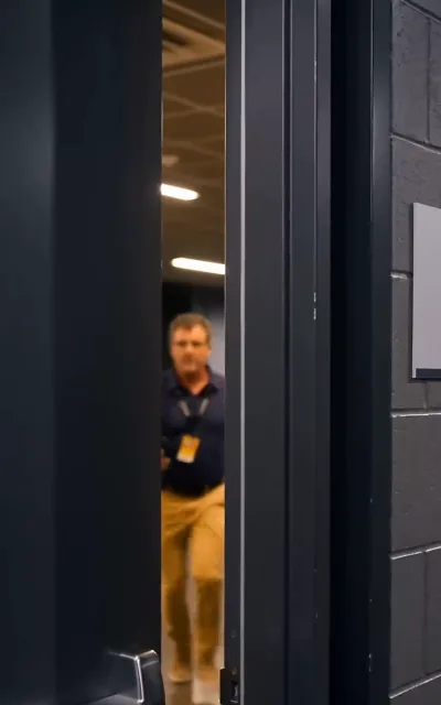 Watched — a middle-aged man in a navy polo and a work lanyard approaching through a door gap: someone is keeping tabs on the champions (ep2; he stays unnamed)