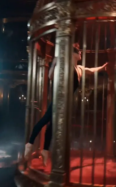 Inside the giant birdcage, the masked dancer bends against the bars in warm red stage light